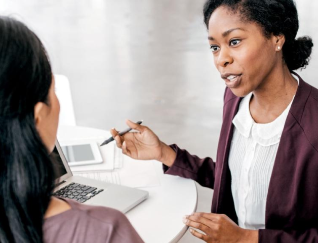 Woman speaking to another woman with pen in hand and computer on table