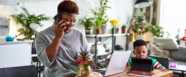 An African American woman sits in kitchen, speaking on mobile phone while in front of laptop, young son sits in background playing on tablet device