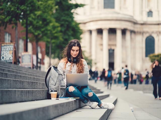 A woman sits on stairs in college setting reviewing finances on laptop