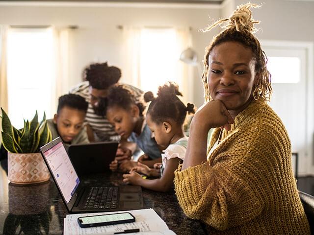 A mother sits at desk with laptop, 4 kids playing on tablet in background