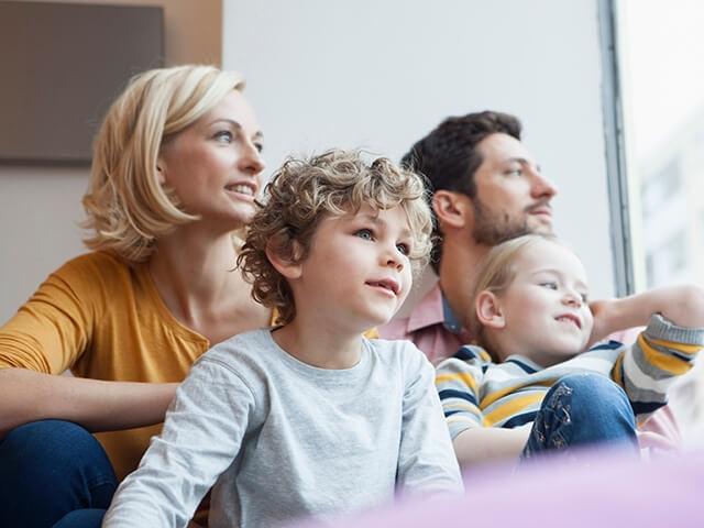 A family of four sits together on couch looking out window