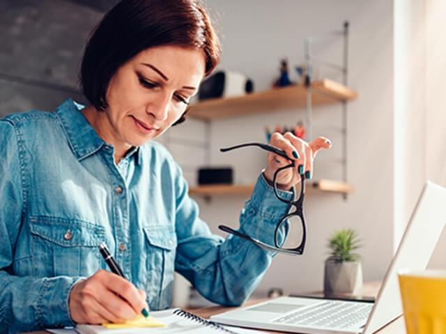 A woman sits at kitchen table with laptop, pen and paper